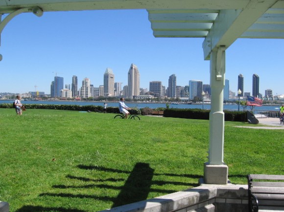 Gazing from a sheltered area with benches across green grass. The skyline of downtown San Diego rises in the background.