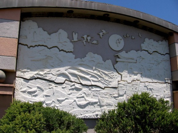 Beautiful artwork on one building's side shows a beach and birds in flight. Swallows have built nests above it near the roof.
