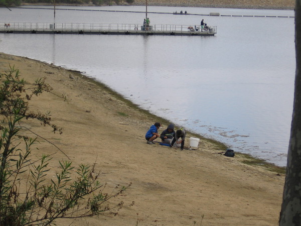 These kids who are fishing huddle together to check out something on the lake's shore.