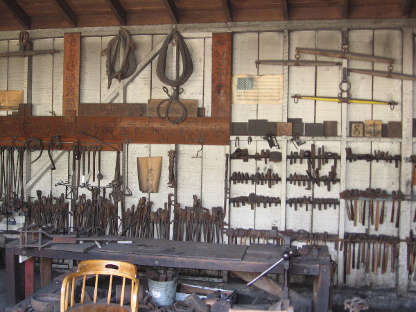 Hammers, bars, wrenches and various blacksmithing tools hang from the rear wall, in addition to harnesses and other items one might find in a livery stable.