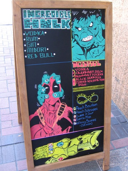 Funny sign on downtown San Diego sidewalk depicts Hulk squeezing Deadpool's head and the Infinity Gauntlet.