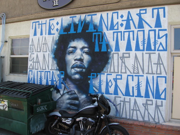 An awesome Jimi Hendrix street mural near dumpster and motorcycle.