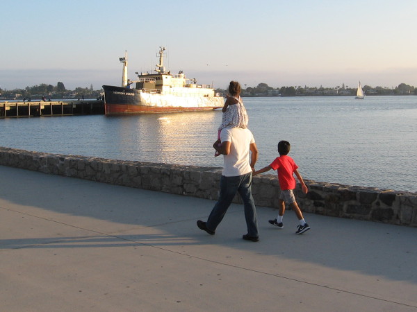A family walks along and gazes out at a sunlit ship.