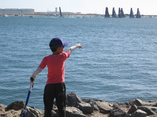 Boy points toward fleet of high-tech foil-equipped catamarans manned by elite level teams. Another wild race is about to begin.