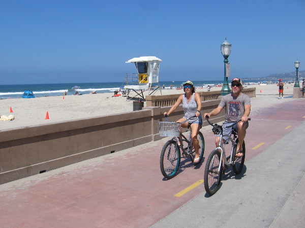 A couple rides bicycles along the Mission Beach boardwalk on a perfect day.