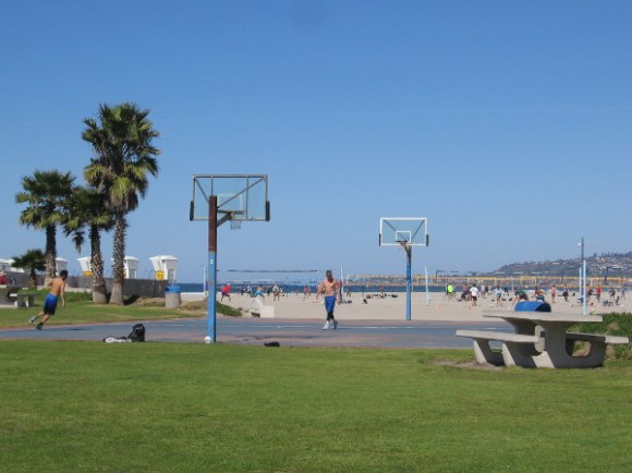 Guys play in the sun at the basketball court.