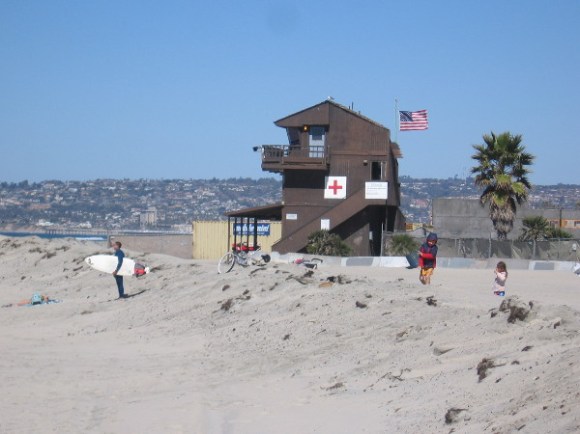 On the beach, a lifeguard building with flag flapping in the sea breeze. Pacific Beach and La Jolla rise to the north.