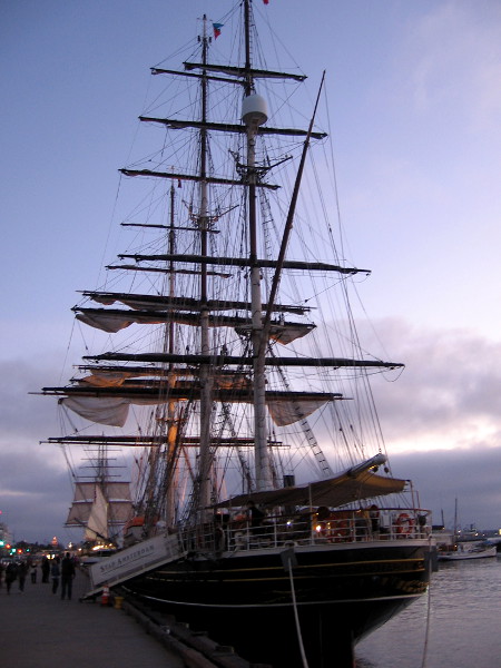 Photo of stern of Stad Amsterdam as the sun is setting behind a bank of clouds. The sails of Star of India, pride of the Maritime Museum of San Diego, can be seen in the distance.