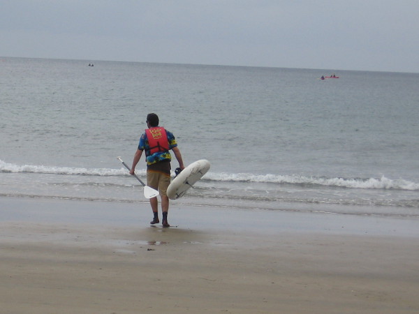 A man carries his kayak across the sand toward the water. It's an overcast winter day, but very pleasant.