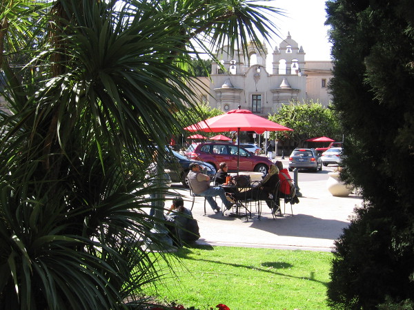 People relax at a table under a red umbrella in the Plaza de Panama. The House of Charm rises in the background.