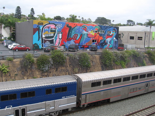An Amtrak Pacific Surfliner train heads south, passing a cool mural titled Myths at Play, just south of the Solana Beach station.