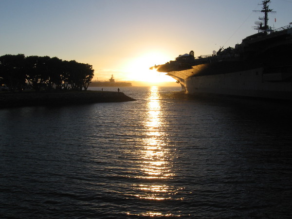 The sun nears the horizon beyond the USS Midway Museum, reflecting from tranquil San Diego Bay.