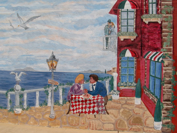 Lovers stand on a balcony, and eat Italian food by the ocean.
