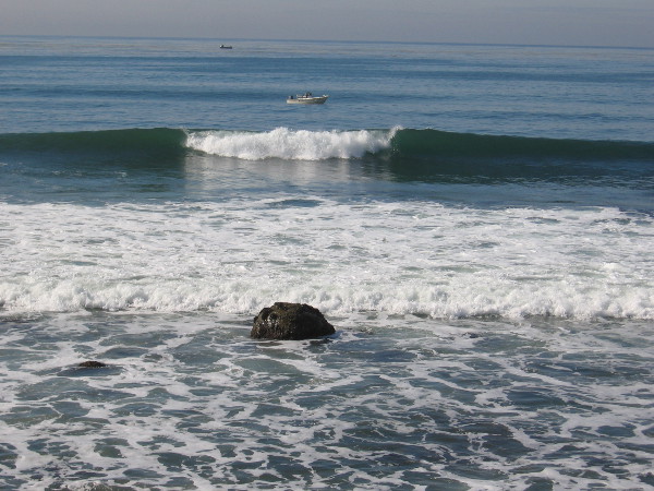 A few rocks stick out of the surf. Fishing boats lie in the water beyond.