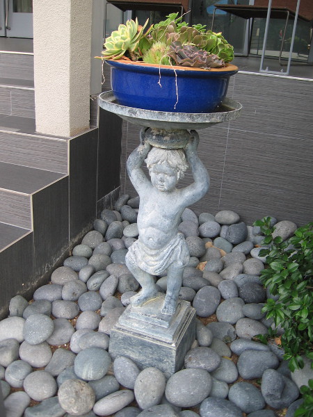 A sculpted cherub balances a potted plant atop its head.