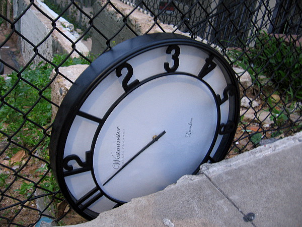 A clock wedged between a sidewalk and fence.