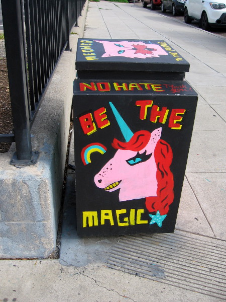 A rainbow and fierce pink unicorn painted on a utility box. BE THE MAGIC
