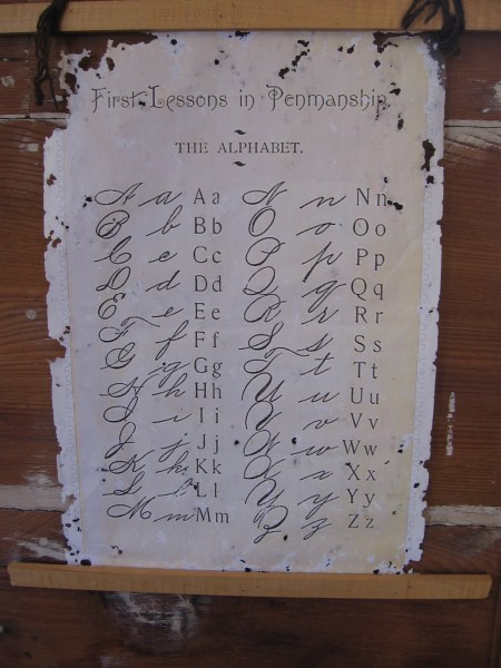 A sheet on the schoolhouse wall contains First Lessons in Penmanship. THE ALPHABET.