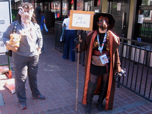 A plague doctor carries a sign: Wash your hands. Can Dr. Beak pry that alien off the face of that nearby patient?